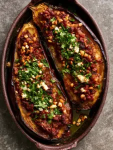 Professional food photography of roasted stuffed eggplant halves with blistered purple skin and golden pine nut garnish. (Stuffed eggplant)